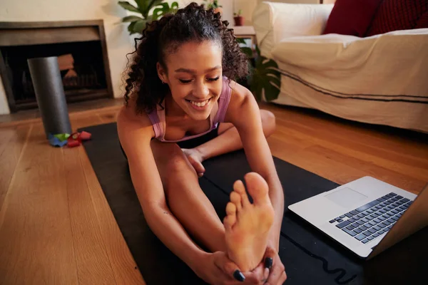 Güzel, mutlu, Afro-Amerikalı genç bir yetişkin kadın dizüstü bilgisayarında yoga egzersiz videosu izlerken oturma odasında esniyor.