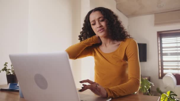 Mujer multicultural obtiene dolor en el cuello mientras trabaja de forma remota desde casa en el ordenador portátil en el mostrador de cocina de moda. — Vídeo de stock