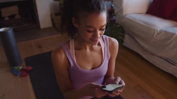 Gelukkig Multi-culturele jonge volwassen vrouw typen op haar telefoon terwijl op yoga mat in moderne lounge thuis — Stockvideo
