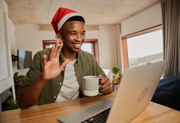 Genç siyah erkek Noel şapkası takıyor, online toplantıda el sallıyor. Mutfak tezgahında dizüstü bilgisayar kullanarak oturmak..