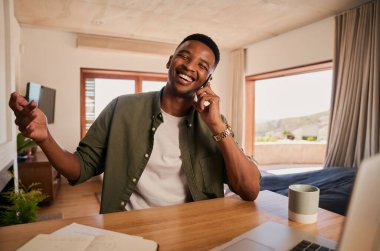 Young adult black male smiling after receiving good news on a phone call. Working from laptop at home in modern apartment.