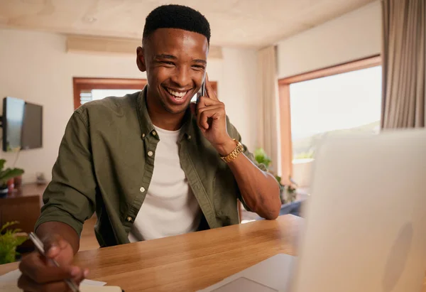 Young adult black male smiling while on phone call with colleague. Remote working from modern home with laptop, notebook and cellphone
