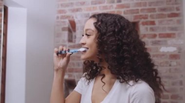 Multicultural young adult female brushing her teeth happily in the modern bathroom mirror. Health and wellness