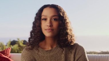 Kameraya bakan çok kültürlü bir kadının portresi. Arkasında güneş ışığı, modern dairenin oturma odasında koltukta oturuyor.