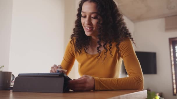 Multicultural young adult female smiling, typing on electronic tablet, working remotely from home at trendy kitchen counter. — Stock video