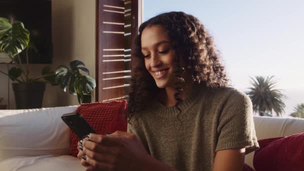 Multiculturele jonge volwassen vrouw glimlacht naar zichzelf terwijl ze een vriend sms 't op haar mobiele telefoon. Zittend op een witte bank in de moderne woonkamer. — Stockvideo