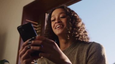 Çok kültürlü genç bir kadın cep telefonuyla yazarken şakaya gülüyor. Modern daire, gündüz vakti boş zaman.