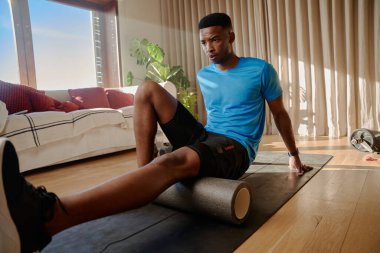 Genç Afro-Amerikan erkek, spor paspası üzerinde köpük bir silindir kullanarak ayak bileğini esnetiyor, evinde, oturma odasında çalışıyor. Yüksek kalite fotoğraf