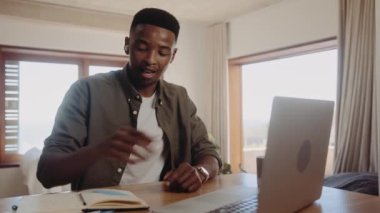 Genç, Afro-Amerikalı zenci bir erkek online bir görüşmede kulaklık takıyor. Bilgisayarını kullanıyor, evden çalışıyor. Günlüğüne not alıyor. Yüksek Kalite 4K video.