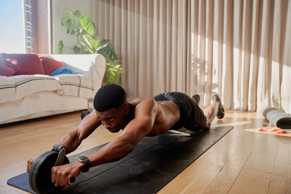 Genç Afro-Amerikan erkek, oturma odasında egzersiz yapan ve yoga minderinin üzerinde egzersiz yapan bir karın tekerleği kullanıyor. Yüksek kalite fotoğraf
