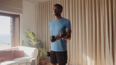 Genç Afro-Amerikan erkek, oturma odasında ağır halterlerle biseps kıvırıyor. Formda ve dayanıklılık eğitimiyle sağlıklı. Yüksek kalite 4K Video