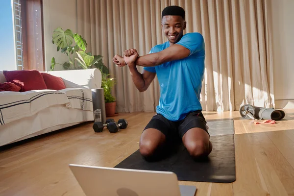 Genç siyahi Afrikalı Amerikalı mutlu ve gülümserken modern dairesinde yoga minderinde online egzersiz yapmadan önce kollarını esnetiyor. Yüksek kalite fotoğraf
