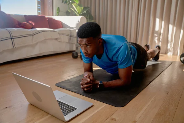 Young African American black male athlete watching workout videos on his laptop doing an online class while holding plank position. Keeping fit, exercising at home. High quality photo