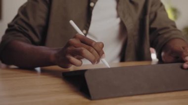 Young adult black African-American male drawing with pen on electronic tablet while working remotely from modern apartment. Graphic designer, digital nomad. 4K Video