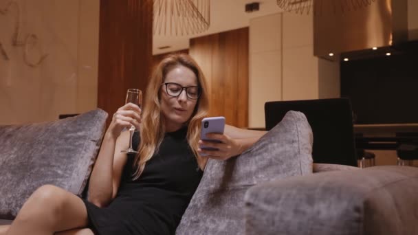 Frau im Abendkleid mit Weinglas lacht auf dem Sofa mit Handy — Stockvideo