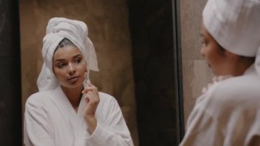 Siyah tenli genç esmer, aynanın önünde bornozlu, yüzüne masaj yapıyor.