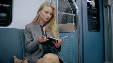 İş sarışını kız metroda oturuyor, deftere yazıyor, kulaklık takıyor.