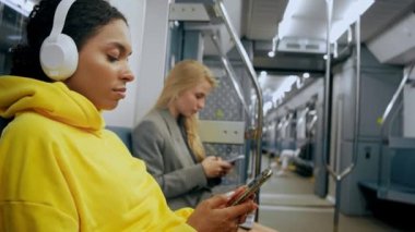 İki genç kız modern metroda oturuyor, telefonlara bakıyor, müzik dinliyorlar.