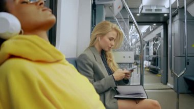 Genç sarışın iş kızı metroda oturuyor ve kulaklıkla telefona bakıyor.
