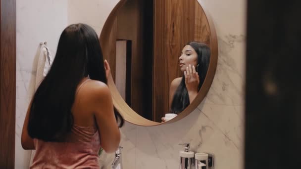 Mladá brunetka s tmavou pletí před zrcadlem jí nanáší krém na obličej. — Stock video