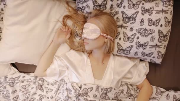 Jong blondje met slaapmasker ligt op bed onder een deken. wakker worden uit de slaap — Stockvideo