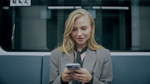 Ung blond flicka sitter i modern tunnelbana, tittar på sin telefon och ler — Stockvideo