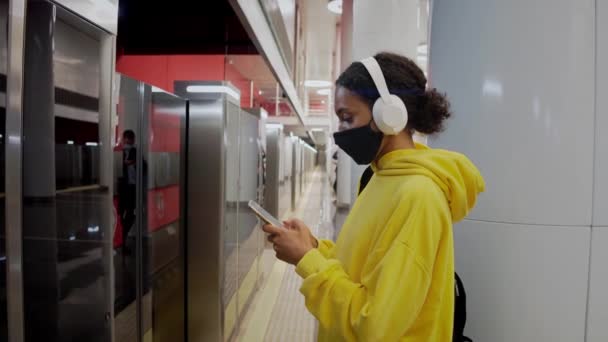 Jeune fille en train d'attendre le métro, portant un masque et un casque, regardant dans le téléphone — Video