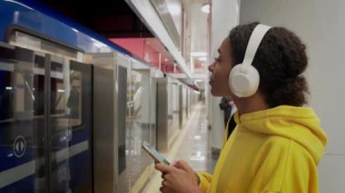 Genç bir kız metro trenine biniyor, kulaklıkla müzik dinliyor ve telefona bakıyor.