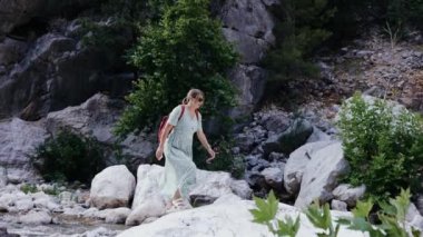 Hamile bir kız sırt çantasıyla bir dağ nehrinin taşları boyunca yürüyor.