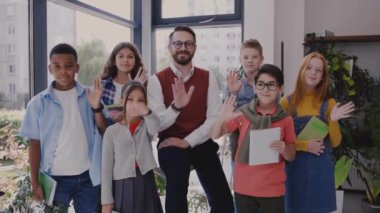 Ortaokul çocukları olan erkek öğretmen kameraya bakıyor ve el sallıyor.