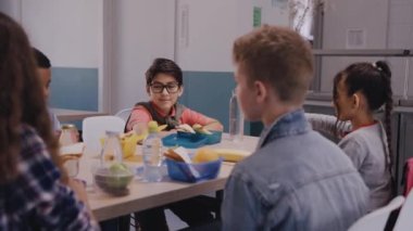 Çocuklar okul kantininde yemek yiyor, konuşuyor ve gülüyorlar.