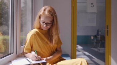 Öğretmen okulun penceresinde oturuyor, deftere yazıyor ve kameraya gülümsüyor.