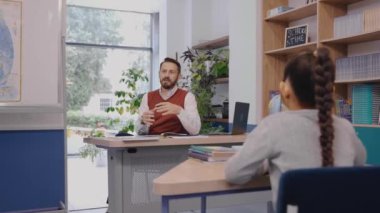 Kız, öğretmenin sorduğu bir soruyu cevaplamak istiyor. Masanın yanında duruyor..