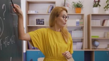 Kadın öğretmen tahtada duruyor ve aktif olarak bir konuyu anlatıyor.