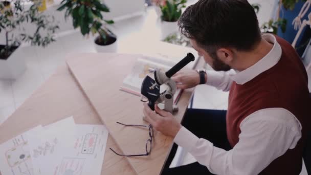 Mann-lærer som bruker mikroskop og ser på biologibok i moderne skole – stockvideo