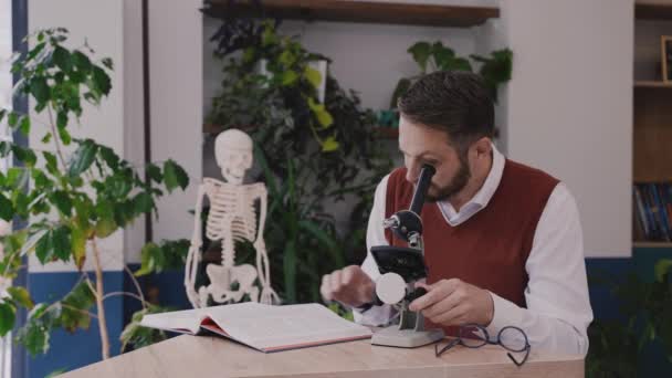 Учитель-мужчина использует микроскоп и просматривает учебник биологии в современной школе — стоковое видео