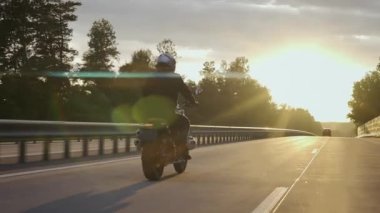 Otoyolda gün batımında ormanda motosiklet süren bir adam var.