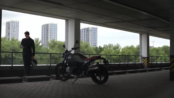 Chauffeur de moto debout sur le parking regardant téléphone, forêt derrière lui — Video