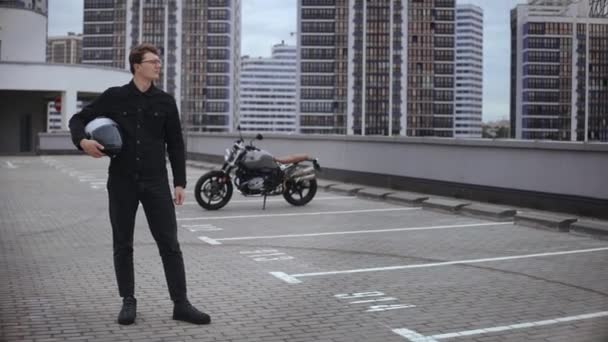 Smart guy stands in front of motorbike with helmet at top of multi-level parking — Stock Video