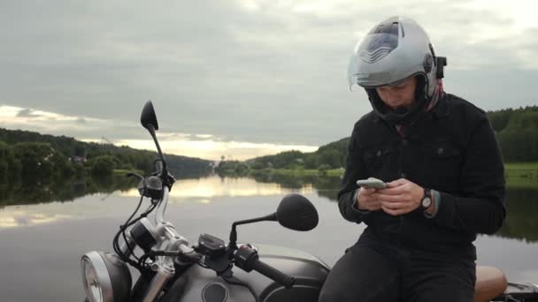 Messagerie du conducteur de moto et regarder son téléphone assis au coucher du soleil — Video