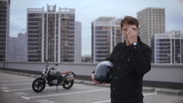 Smart kille står framför motorcykel med hjälm på toppen av flera nivåer parkering — Stockvideo