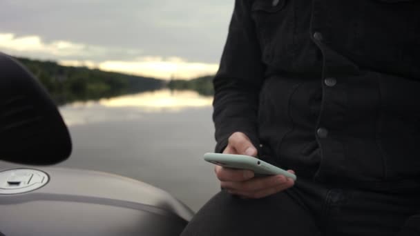 Foto ravvicinata di un uomo che messaggia al telefono seduto sulla moto vicino al lago — Video Stock