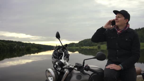 Man talar i telefonen sitter på scrambler motorcykel vid sjön — Stockvideo