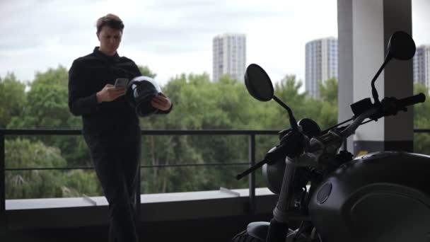 Motorista de moto de pé no estacionamento olhando para o telefone, floresta atrás dele — Vídeo de Stock
