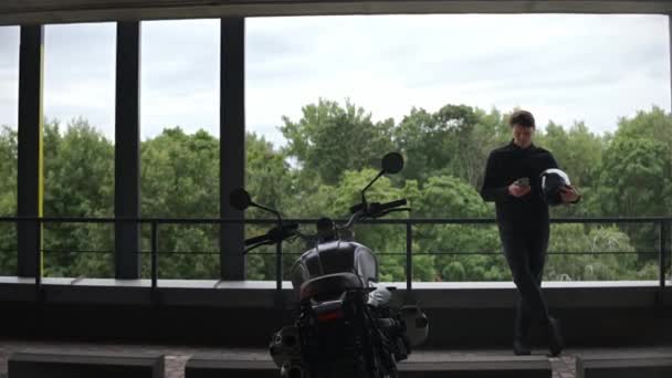 Chauffeur de moto debout sur le parking regardant téléphone, forêt derrière lui — Video