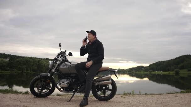 Motociclista che parla al telefono seduto su uno scrambler in riva al lago in autunno — Video Stock
