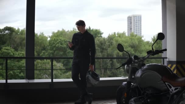 Chauffeur de moto debout sur le parking regardant téléphone, forêt derrière lui — Video