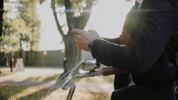 Чоловік у костюмі дивиться на телефон, спираючись на велосипед у парку, крупним планом — стокове відео