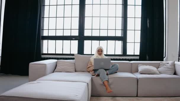 Ung muslim flicka sitter på soffan tittar på laptop i ljusa beige studio — Stockvideo