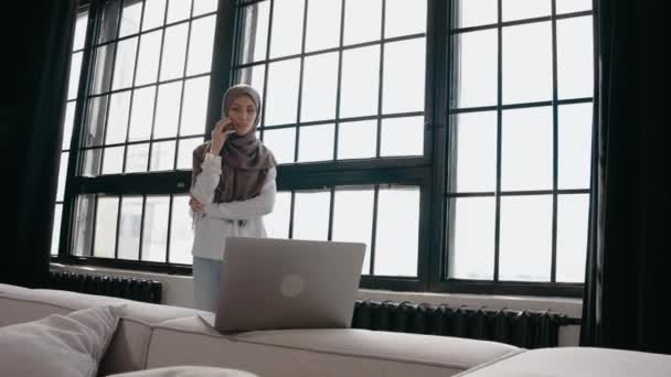 Muselman flicka talar på telefon och tittar på laptop, leende i ljus studio — Stockvideo